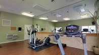 This single-floor fitness center offers a clean and functional environment tailored for cardio and general exercise. The space is outfitted with a selection of cardio equipment, including treadmills and elliptical trainers, positioned to allow comfortable use. A full-length mirror spans a large portion of one wall, visually expanding the room and allowing users to monitor their form during workouts. The flooring is a warm hardwood that adds a polished, welcoming feel underfoot, while rubber mats beneath the equipment provide stability and floor protection. The walls are painted in a soft sage green, contributing to a calm and energizing atmosphere. Overhead, recessed flush-mount lighting panels provide consistent, even illumination throughout the space. A tiled wainscoting detail runs along the lower portion of the walls, adding a subtle architectural accent. A bulletin board is mounted near the entrance area, likely used for community notices or fitness information. Ventilation is supported by ceiling vents, ensuring airflow during workouts. Overall, the space strikes a balance between practicality and comfort, making it a solid and inviting facility for everyday fitness activity.