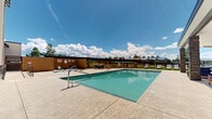 Located at 1002 N Fourth St in Flagstaff, Arizona, this outdoor amenity space is designed to offer a resort-style experience beneath the wide Arizona sky. At its centerpiece sits a generously sized swimming pool with crystal-clear blue water, framed by a broad concrete pool deck ideal for lounging and socializing. A dedicated spa or wading area is positioned adjacent to the main pool, providing an additional relaxation option.

A covered cabana pavilion anchors one side of the pool deck, supported by handsome stone-clad columns beneath a wide, sheltering roof. The shaded area is furnished with plush seating arrangements, creating a comfortable outdoor living room for gatherings. Directly beside it, a well-equipped outdoor kitchen and bar area features built-in grills and high-top seating, perfect for entertaining.

On the opposite side of the pool, a lush artificial turf lawn is lined with modern sling-style chaise lounges and accent tables, offering a vibrant green setting for sunbathing. The surrounding multi-story residential buildings, finished in a contemporary mix of stone, wood-look siding, and white paneling, frame the space and contribute to a cohesive community atmosphere. Mature pines and open views of the Flagstaff sky complete the inviting, mountain-town ambiance of this outdoor retreat.