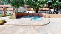 This outdoor amenity area is centered around a circular spa/hot tub with light turquoise water, tiled in dark mosaic along its rim and set into a broad, stone-tiled deck. The deck provides generous space for lounging and is furnished with red metal chairs arranged around the spa, offering residents comfortable seating in a communal setting. Mature shade trees frame the area, providing natural canopy and softening the sun exposure during warmer months.

The spa area is enclosed by a low stucco perimeter wall finished in a warm earth tone, consistent with the architectural style of the surrounding multi-story residential buildings. These buildings feature tan stucco exteriors, red clay tile rooflines, and balconies with metal railings, presenting a cohesive Southwestern aesthetic throughout the complex.

Beyond the spa courtyard, views open to a separate swimming pool area, visible through iron fencing and lush landscaping that includes trimmed hedges, shrubs, and tall palm trees. The grounds are well-maintained with green lawns and planted beds adding color and texture. A utility/amenity building adjacent to the spa provides access to community facilities. Overall, the space offers a relaxed, resort-influenced outdoor environment suited for both leisure and social activity.