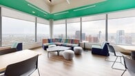 Located at 10 Bay St S in downtown Hamilton, Ontario, this dynamic single-floor workspace offers a thoughtfully designed open-plan environment ideal for collaboration and productivity. The space is defined by its striking interior aesthetic, featuring a bold green ceiling accent that anchors the layout and vivid, geometric mural walls in blue, purple, and yellow that inject energy and personality throughout.

Floor-to-ceiling windows line the perimeter, flooding the space with abundant natural light and offering panoramic views of the Hamilton skyline and beyond — a standout feature that lends an elevated, airy atmosphere to the entire floor. Light wood-tone flooring runs throughout, complementing the bright, contemporary palette.

The layout supports a variety of work styles, with open hot-desking areas furnished with collaborative tables and modern chairs, as well as comfortable lounge seating featuring colorful upholstered sofas and soft ottomans positioned to take full advantage of the sweeping views. A kitchenette area with bar-height seating and a built-in counter provides a social hub for breaks and informal gatherings. Glass-walled meeting rooms offer semi-private areas for focused work or small group discussions.

Overhead, geometric acoustic ceiling panels and sculptural angular lighting fixtures add architectural interest while supporting acoustics in the open setting. Exposed concrete columns punctuate the space, adding an industrial character that balances the vibrancy of the décor.