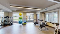 This contemporary fitness studio is thoughtfully designed to support a wide range of exercise activities within a single, open-plan floor. The space features light wood-toned flooring throughout, lending a clean and inviting aesthetic that complements the neutral warm-toned walls and modern recessed linear lighting overhead.

A row of Spinning brand stationary bikes lines one side of the studio, providing dedicated space for cycling workouts. The opposite end of the room is anchored by a large wall-mounted flat-screen display, ideal for instructor-led or virtual classes. Full-length mirrors span an entire wall, enhancing the sense of openness and allowing participants to monitor their form during exercise.

A curated collection of color-coded exercise balls in varying sizes is neatly organized on a vertical rack, adding a vibrant visual element to the space. Step platforms are also available, supporting aerobic and strength training exercises. Additional fitness equipment, including resistance and strength training apparatus, is accessible along the perimeter.

Generous floor-to-ceiling windows on multiple walls allow abundant natural light to fill the room and offer views of the surrounding streetscape, creating an energizing atmosphere. The overall design strikes a balance between functional fitness infrastructure and a welcoming, well-lit environment suited for group or individual training sessions.