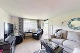 This single-floor home offers a thoughtfully arranged layout encompassing a living room, dining room, kitchen, bedroom, and bathroom. The living room is bright and inviting, featuring large windows dressed with patterned curtains that allow generous natural light to fill the carpeted space. A glass-paneled door provides direct access to an outdoor patio, enhancing the connection to the surrounding greenery visible outside. The dining room, situated adjacent to the kitchen, features wood-look flooring, a ceiling fan with light fixture, and a wide window overlooking a lush outdoor setting, creating a pleasant atmosphere for meals. The galley-style kitchen is equipped with warm wood cabinetry, dark granite-style countertops, a double-basin sink, and a full suite of appliances, offering efficient and practical use of space. A pass-through window connects the kitchen to the dining area. The bedroom provides a comfortable retreat with plush carpet, sheer curtains, and a reach-in closet offering ample storage. The bathroom is clean and bright, featuring white subway tile, a single-sink vanity with white cabinetry, and a large mirror with overhead lighting. An exterior covered pavilion structure and green landscaping are visible in the outdoor area, adding to the overall appeal of the property.