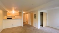This single-floor residential unit offers a practical and functional open-concept layout designed for comfortable everyday living. The main living area is finished with wall-to-wall carpeting that adds warmth underfoot and flows seamlessly throughout the space. A raised kitchen counter serves as a natural divider between the living area and the kitchen, which features light wood cabinetry and recessed lighting that casts a warm, inviting glow over the workspace. A structural column near the kitchen adds a subtle architectural detail to the open floor plan. Along the back wall, a hallway leads to additional rooms, with multiple doors providing access to separate spaces within the unit. An electrical panel is conveniently mounted on the wall, and a floor-level vent indicates a central heating and cooling system. To the right, an open doorway reveals a spacious walk-in closet complete with a clothing rod, offering generous storage capacity. The neutral white and cream wall palette throughout the space creates a clean, bright atmosphere that makes the interior feel open and easily customizable. Overall, the layout is straightforward and efficient, making good use of every area within the single-floor footprint.
