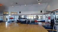 Located at 5051 Open Meadow Dr in Columbus, Ohio, this single-floor fitness facility offers a well-equipped and open workout environment designed to support a wide range of exercise needs. The main workout area features soaring ceilings that contribute to an airy, expansive atmosphere, complemented by large windows that allow generous natural light to fill the space. A full wall of mirrors runs along one side of the room, visually enlarging the area and providing exercisers with clear sightlines for form and technique. The hardwood flooring transitions to rubber mat sections beneath the weight equipment, offering both aesthetic warmth and functional safety. The facility is outfitted with a variety of free weights, including a full dumbbell rack, barbells, and weight benches, alongside plate-loaded strength machines to accommodate both beginner and advanced training. Track lighting mounted on the ceiling provides focused illumination throughout, while a ceiling fan supports air circulation during intense workouts. Decorative red and black accent striping along the upper walls adds a bold, athletic character to the overall design. The layout is thoughtfully organized, providing ample open floor space for stretching, bodyweight exercises, or group fitness activity alongside the dedicated strength training zones.