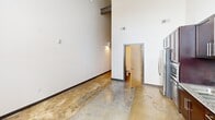 Located at 144 E Fourth St in Burlington, NC, this single-floor studio offers an industrial-modern aesthetic defined by dramatically high ceilings and polished concrete flooring throughout. The open main area showcases exposed brick accents alongside crisp white walls, creating a striking contrast. The well-appointed kitchen features rich dark wood cabinetry, granite countertops, and a full suite of stainless steel appliances including a refrigerator with ice maker, range, microwave, and dishwasher. A dedicated laundry area houses a full-size washer and dryer with wire shelving above. The bathroom includes a combination tub and shower with chrome grab bars.