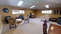 Located at 4700 176th St SW in Lynwood, Washington, this single-floor space offers a well-appointed and versatile layout designed for both comfort and functionality. The interior is characterized by vaulted ceilings adorned with multiple skylights that flood the space with natural light, complemented by recessed lighting throughout.

The main open area is thoughtfully divided into distinct functional zones. A lounge area features patterned accent chairs, an ottoman, and a sofa arranged around a central rug, creating an inviting gathering spot. Adjacent to this is a dedicated workspace with a desk and computer setup, ideal for productivity. A separate media area includes a wall-mounted flat-screen television, a coffee table, and additional seating including armchairs and a sofa, providing a relaxed entertainment zone.

The kitchenette is equipped with dark-stained cabinetry, granite-style countertops, a stainless steel sink, a microwave, and a coffee maker — offering convenient refreshment options without a full kitchen footprint. A staircase visible from the lounge area connects to the lower level of the building.

Warm neutral tones on the walls, plush carpeting underfoot, and windows fitted with wood blinds contribute to a cohesive, welcoming ambiance throughout the space.