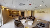 This well-appointed single-floor amenity space is thoughtfully designed to serve as a comfortable and functional gathering area. The main lounge is anchored by a generous seating arrangement featuring dark charcoal armchairs and sofas paired with sleek white round coffee tables and elegant gold accent side tables, all set atop a neutral-toned area rug. A decorative metallic wall sculpture adds an artistic focal point to the warm, neutral-painted walls, while recessed lighting throughout maintains a welcoming ambiance. Large floor-to-ceiling windows along one wall flood the space with natural light and offer pleasant views of the surrounding green landscape, enhanced by wall-mounted sconce lighting on either side. A wall-mounted flat-screen television provides additional entertainment options in the lounge area. The kitchenette, positioned at one end of the space, is both practical and visually striking. It features a long white countertop, dark wood cabinetry, a double stainless steel sink, a smooth-top electric range with a stainless steel range hood, a dishwasher, and a full-size refrigerator. The kitchenette's standout feature is an expansive textured accent wall in deep charcoal and gold tones, adding dramatic visual interest. Light tile flooring in this zone contrasts beautifully with the lounge's carpeted area, clearly delineating the two functional spaces.