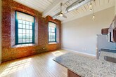 Unit 326 at 1082 RT-79 in Fall River, MA is a one-bedroom, two-bathroom loft-style residence that blends industrial character with modern finishes. The open-plan living and kitchen area immediately impresses with soaring ceilings accented by exposed ductwork, a white-painted wood-beam ceiling, and a ceiling fan. Warm hardwood flooring runs throughout the main living space, filling the room with natural light that pours in through oversized arched windows framed by original exposed brick walls — a striking nod to the building's historic mill architecture. The kitchen is equipped with dark espresso cabinetry, granite countertops, a stainless steel dishwasher, a peninsula island for casual dining, and pendant lighting overhead. The generously sized primary bedroom continues the hardwood flooring and neutral toned walls. The hallway connects to two bathrooms, both featuring large-format tile flooring in a soft gray tone. One bathroom includes a full soaking tub and shower combination with clean white surrounds, while the other offers vanity storage with granite countertops and built-in closet space. The overall ambiance is one of urban sophistication rooted in historic New England mill charm, making this a distinctive and functional living space.