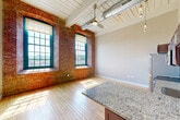 Unit 326 at 1082 RT-79 in Fall River, MA is a one-bedroom, two-bathroom loft-style residence that blends industrial character with modern finishes. The open-plan living and kitchen area immediately impresses with soaring ceilings accented by exposed ductwork, a white-painted wood-beam ceiling, and a ceiling fan. Warm hardwood flooring runs throughout the main living space, filling the room with natural light that pours in through oversized arched windows framed by original exposed brick walls — a striking nod to the building's historic mill architecture. The kitchen is equipped with dark espresso cabinetry, granite countertops, a stainless steel dishwasher, a peninsula island for casual dining, and pendant lighting overhead. The generously sized primary bedroom continues the hardwood flooring and neutral toned walls. The hallway connects to two bathrooms, both featuring large-format tile flooring in a soft gray tone. One bathroom includes a full soaking tub and shower combination with clean white surrounds, while the other offers vanity storage with granite countertops and built-in closet space. The overall ambiance is one of urban sophistication rooted in historic New England mill charm, making this a distinctive and functional living space.