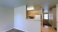 This single-story space is designed with an open and functional layout that promotes a connected flow between the kitchen and living areas. The kitchen is positioned behind a half-wall that doubles as a pass-through counter, finished with a light granite-style surface ideal for casual dining or food preparation. Light wood cabinetry lines the upper walls of the kitchen, providing ample storage, while a range with a built-in overhead exhaust hood and a full-size refrigerator offer essential cooking amenities. Overhead fluorescent lighting ensures the kitchen remains well-lit. The surrounding walls are finished in neutral tones, contributing to a clean and understated aesthetic throughout. Carpeted flooring in the adjacent living area adds a layer of warmth and comfort to the space. A window visible in the background allows natural light to filter into the living area, enhancing the overall brightness of the unit. The architectural use of a partial dividing wall creates a sense of defined zones while maintaining an open, airy feel across the shared living and kitchen space.