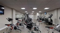 This fitness center offers a comprehensive workout environment housed within a single-floor layout. The space features a drop ceiling with evenly spaced recessed fluorescent panels that provide consistent, bright illumination throughout the room. Neutral grey walls and light-toned flooring create a clean, functional atmosphere conducive to exercise.

The facility is well-stocked with a variety of equipment, including a row of stationary spin bikes along one side, ideal for cycling workouts. A multi-function cable and weight machine occupies a central position, offering versatility for strength training exercises. On the opposite side, a line of elliptical trainers and treadmills provides ample options for cardiovascular workouts. A wall-mounted television is positioned toward the far end of the room, allowing users to stay entertained during their sessions.

Large mirrors line one wall, visually expanding the space and allowing users to monitor their form during exercise. The room provides enough floor space to navigate comfortably between equipment. Two doors are visible at the far end, offering convenient access points. Overall, the fitness center presents a practical and organized environment suitable for a range of fitness routines.