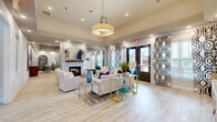 Located at Seven Oaks Boulevard in Smyrna, Tennessee, this elegantly appointed community clubhouse offers a thoughtfully designed, open-concept gathering space that seamlessly blends comfort and contemporary style. The single-floor layout provides a generous, flowing footprint that accommodates multiple distinct lounge and social areas.

At the heart of the space stands a striking floor-to-ceiling white stone fireplace, complemented by a mounted flat-screen television, serving as a dramatic architectural focal point. Multiple seating arrangements — including plush neutral sofas, navy accent chairs, and leather lounge seats — are positioned throughout, creating both intimate conversation areas and open social zones.

Light-toned wood-look flooring runs throughout, enhancing the bright, airy ambiance generated by recessed lighting and a statement gold pendant fixture overhead. Bold black-and-white geometric drapery panels add a graphic design element that contrasts beautifully with the neutral color palette.

A curved wet bar with stone-clad detailing and bar seating offers a dedicated refreshment area, while a separate dining nook provides additional functionality. Large windows and glass entry doors invite abundant natural light and offer views to the outdoors. Curated artwork, decorative plants, and layered area rugs complete the polished, welcoming atmosphere of this versatile community space.