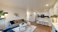 This thoughtfully designed single-floor residence offers a seamless blend of modern aesthetics and functional living. The open-concept layout connects the living area and kitchen, creating a bright and welcoming atmosphere enhanced by recessed lighting and natural light streaming through large windows.

The living area is generously proportioned, featuring warm hardwood-style flooring, neutral white walls, and ample space for comfortable seating arrangements. A large window floods the room with natural light, contributing to the airy feel of the space.

The kitchen is well-equipped with stainless steel appliances, including a refrigerator, range, microwave, and dishwasher. White upper cabinetry pairs with wood-toned lower cabinets and light quartz countertops, while a tasteful tile backsplash adds texture. A kitchen island with bar seating and a stainless steel sink enhances both functionality and social appeal. Double pantry doors provide generous storage.

The bedroom is a calm retreat with plush carpeting, crisp white walls, and a bright window. The bathroom features a wide vanity with quartz countertops and an extended mirror, complemented by convenient in-unit stacked washer and dryer. A built-in linen closet with shelving adds practical storage within the bathroom space.