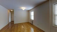 This single-floor space presents a clean, understated interior highlighted by warm-toned hardwood floors that run throughout, adding a sense of continuity and warmth to the layout. The main room is an open, well-proportioned area with smooth white walls and white trim baseboards that lend a crisp, neutral backdrop adaptable to a variety of furnishing styles. A single window allows natural light to filter into the room, contributing a bright and airy quality to the space. Beneath the window sits a traditional cast-iron radiator, indicating a classic heating system suited for cooler climates. The ceiling is smooth and finished with a simple flush-mount light fixture that provides ambient overhead illumination. A doorway visible along one wall suggests connectivity to additional areas of the unit, hinting at a practical, straightforward floor plan. The overall ambiance is minimal and move-in ready, offering a functional and timeless aesthetic that serves as a versatile foundation for personalization.
