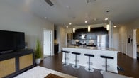 This sleek, single-floor residence showcases a thoughtfully designed open-concept layout that seamlessly connects the kitchen and living areas. The kitchen is a standout feature, outfitted with rich dark cabinetry that extends to the upper walls, providing generous storage. Stainless steel appliances, including a refrigerator, stove, and microwave, add a polished, professional quality to the space. A large kitchen island with a light granite countertop serves as both a functional workspace and a social hub, complemented by a row of modern black and chrome bar stools. Pendant lighting with warm-toned cylindrical shades hangs above the island, adding an elegant focal point while enhancing the overall ambiance. Recessed lighting throughout ensures the space remains bright and well-lit. The rich dark hardwood flooring grounds the interior with warmth and continuity, flowing through the open living area where a cozy fireplace and a lush potted plant introduce natural and inviting elements. The neutral-toned walls and white area rug soften the contrast of the darker finishes, creating a balanced, contemporary aesthetic. A hallway leads to additional rooms, hinting at a well-organized and functional floor plan throughout.