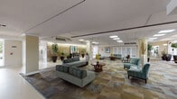 This single-floor space presents a thoughtfully designed lobby and common area that exudes a warm, hospitality-inspired ambiance. Upon entry, visitors are greeted by a bright, open foyer with terrazzo-style flooring that transitions into richly patterned carpeting across the main lounge areas. A standout feature is the dramatic accent wall clad in an earth-toned stone or metallic mural finish, flanked by tall, dark-potted tropical plants that add a lush, organic element to the interior.

The concierge desk serves as a central hub, clearly marked and positioned to assist residents and guests with ease. Adjacent seating areas are furnished with teal and neutral-toned sofas, accent chairs, and warm wood coffee tables, creating multiple intimate conversation zones throughout the open floor plan.

Natural light floods the space through floor-to-ceiling glazed windows along one wing, offering views of manicured outdoor landscaping with palm trees and tropical foliage. Pendant lighting and recessed skylight panels supplement the natural light, maintaining a bright and airy atmosphere throughout the day.

Artwork and decorative accessories are thoughtfully placed, adding color and personality to the neutral wall tones. The elevator corridor and connecting hallways are clean and well-lit, ensuring smooth circulation throughout the space. Overall, the design strikes a balance between refined elegance and comfortable functionality.