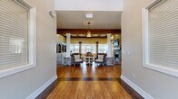 Located at 2880 Commonwealth Drive in Spring Hill, Tennessee, this single-floor space offers a generously sized open-concept layout that seamlessly integrates a full kitchen and expansive living area. Rich hardwood flooring flows throughout, anchoring the warm, rustic aesthetic that defines the space.

The living area is a standout, characterized by soaring vaulted ceilings adorned with bold, exposed wood beams that add architectural drama and a sense of grandeur. White decorative columns with stone bases divide the space into distinct seating zones without sacrificing the open feel. A floor-to-ceiling stone fireplace serves as a striking focal point, while built-in display shelving along the walls provides both function and character. Multiple windows flood the room with natural light and offer views of the surrounding neighborhood.

The kitchen is equally impressive, featuring cream cabinetry, a tiled backsplash, stainless steel appliances, and a spacious central island with bar seating and open shelving. Exposed wood beams carry through from the living area, maintaining design continuity. Industrial-style pendant lights hang above the island, adding a modern farmhouse touch. A sliding barn door accent further enhances the rustic charm. A glass-panel exterior door provides convenient access to the outdoors, reinforcing the bright, welcoming ambiance throughout.