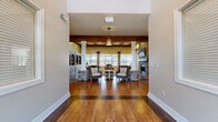 Located at 2880 Commonwealth Drive in Spring Hill, Tennessee, this single-floor space offers a generously sized open-concept layout that seamlessly integrates a full kitchen and expansive living area. Rich hardwood flooring flows throughout, anchoring the warm, rustic aesthetic that defines the space.

The living area is a standout, characterized by soaring vaulted ceilings adorned with bold, exposed wood beams that add architectural drama and a sense of grandeur. White decorative columns with stone bases divide the space into distinct seating zones without sacrificing the open feel. A floor-to-ceiling stone fireplace serves as a striking focal point, while built-in display shelving along the walls provides both function and character. Multiple windows flood the room with natural light and offer views of the surrounding neighborhood.

The kitchen is equally impressive, featuring cream cabinetry, a tiled backsplash, stainless steel appliances, and a spacious central island with bar seating and open shelving. Exposed wood beams carry through from the living area, maintaining design continuity. Industrial-style pendant lights hang above the island, adding a modern farmhouse touch. A sliding barn door accent further enhances the rustic charm. A glass-panel exterior door provides convenient access to the outdoors, reinforcing the bright, welcoming ambiance throughout.
