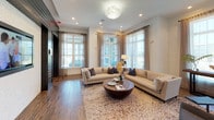 Located at 1605 Barclay Point Blvd in Wilmington, North Carolina, this single-floor space showcases a thoughtfully designed interior that blends contemporary coastal aesthetics with warm, inviting details. The layout is defined by a striking contrast of crisp white shiplap walls and bold dark wood paneling, creating a visually dynamic atmosphere throughout.

The main lounge area serves as the centerpiece of the space, featuring two generously sized neutral-toned sofas arranged around a sculptural round coffee table atop a richly patterned area rug. Recessed lighting, a decorative pendant fixture, and abundant natural light from large multi-pane windows draped with sheer and linen curtains contribute to an open, airy ambiance. Views to the exterior are visible through floor-to-ceiling glass-paneled doors and oversized windows.

A secondary seating nook offers a more intimate setting, with soft sofas, layered accent pillows, and a side lamp, ideal for quiet conversation or relaxation. Rich hardwood-style flooring flows consistently throughout, anchoring the various zones together.

A corridor lined with dark shiplap-clad doorways connects the main areas, adding architectural depth and a sense of purposeful flow. Distinctive decorative elements, including sculptural accents and unique wall textures, add character and personality to this well-appointed communal space.