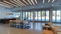 This single-floor space showcases a clean, contemporary design defined by an expansive wall of floor-to-ceiling glass panels and folding doors that span nearly the entire exterior-facing side of the room. These large-format glazed openings flood the interior with abundant natural light while providing unobstructed views of an outdoor pool and surrounding tree-lined grounds, creating a seamless visual connection between indoor and outdoor environments.

The ceiling is a notable architectural feature, combining a warm wood-panel finish with recessed linear LED lighting strips arranged in a parallel pattern, adding both texture and a modern aesthetic to the overhead plane. Polished concrete-style flooring in a neutral tone runs throughout, contributing to the open, airy feel of the space.

The layout is divided into distinct functional zones. A communal dining or work area is anchored by a substantial rectangular wood table surrounded by modern chairs, suitable for group gatherings or collaborative use. A separate lounge area to one side features upholstered seating with patterned cushions, offering a more relaxed setting. The overall ambiance is bright, welcoming, and versatile, well-suited for both social and productive activities.