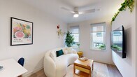 Located at 2009 N Howard St in Philadelphia, PA, this efficiently designed one-bedroom, one-bathroom residence makes smart use of its compact footprint across a single floor. The open-plan living room and kitchen form the heart of the unit, bathed in natural light from multiple windows fitted with roller shades. The living area features light hardwood-style flooring, a ceiling fan, and a wall-mounted flat-screen TV, creating a comfortable and functional gathering space. The kitchen is thoughtfully appointed with stainless steel appliances—including a range, dishwasher, microwave, and refrigerator—complemented by white cabinetry, open shelving with a tile backsplash, and a compact dining area. The bedroom offers a ceiling fan, a built-in wardrobe with overhead storage, and a window with exterior views, providing a cozy and organized retreat. A stacked washer and dryer unit is conveniently tucked behind louvered doors within the bedroom. The full bathroom features large-format grey tile flooring, a sleek floating vanity with an illuminated mirror, and a separate shower enclosure. Throughout the unit, crisp white walls, consistent flooring, and modern black hardware fixtures create a cohesive, contemporary aesthetic ideal for urban living in Philadelphia.