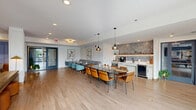 This well-appointed amenity lounge spans a generous single floor, offering a thoughtfully designed multi-zone layout that balances social gathering and relaxed recreation. Light hardwood flooring runs throughout, unifying the space while complementing its contemporary aesthetic.

The dining and kitchenette area anchors one end of the space, featuring a large communal table surrounded by a mix of teal and amber chairs, pendant lighting overhead, and a banquette along the wall. The kitchenette is outfitted with white cabinetry, a textured stone tile backsplash, open shelving with decorative accents, a beverage refrigerator, and a coffee station.

A set of glass-paneled double doors provides entry and connects the lounge to adjoining amenity spaces, including what appears to be a fitness area. Natural light filters in through these doors, brightening the open circulation zone.

The lounge area offers a striking double-sided fireplace set within a wood-and-stone bookshelf partition, creating a natural divide between zones. Built-in shelving with blue accent walls, a wall-mounted television, a pool table with a brass pendant fixture, and a mix of seating arrangements—including a sofa, lounge chairs, and ottomans—complete the space with a warm, mid-century modern sensibility.