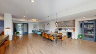 This well-appointed amenity lounge spans a generous single floor, offering a thoughtfully designed multi-zone layout that balances social gathering and relaxed recreation. Light hardwood flooring runs throughout, unifying the space while complementing its contemporary aesthetic.

The dining and kitchenette area anchors one end of the space, featuring a large communal table surrounded by a mix of teal and amber chairs, pendant lighting overhead, and a banquette along the wall. The kitchenette is outfitted with white cabinetry, a textured stone tile backsplash, open shelving with decorative accents, a beverage refrigerator, and a coffee station.

A set of glass-paneled double doors provides entry and connects the lounge to adjoining amenity spaces, including what appears to be a fitness area. Natural light filters in through these doors, brightening the open circulation zone.

The lounge area offers a striking double-sided fireplace set within a wood-and-stone bookshelf partition, creating a natural divide between zones. Built-in shelving with blue accent walls, a wall-mounted television, a pool table with a brass pendant fixture, and a mix of seating arrangements—including a sofa, lounge chairs, and ottomans—complete the space with a warm, mid-century modern sensibility.