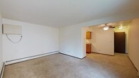 This single-floor space presents a clean, functional layout with an open feel throughout. The main living area is finished with neutral wall-to-wall carpeting and bright white walls, creating a simple, versatile backdrop. A baseboard heater runs along one wall, providing a practical heating solution. The space transitions seamlessly into an adjacent kitchen area, visible through an open passageway. The kitchen features warm-toned wood cabinetry along the upper and lower sections, offering ample storage. A ceiling fan is installed in the kitchen area, promoting air circulation throughout the connected spaces. The white walls and light-colored flooring contribute to a bright, airy atmosphere, while the open sightlines between the living and kitchen areas enhance the overall sense of space. The layout is straightforward and practical, well-suited for everyday living. Dark baseboard trim provides a subtle contrast against the light walls and carpet, adding a modest finishing detail throughout the interior.