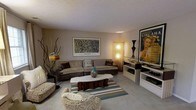 This single-floor space showcases a thoughtfully designed living area that blends mid-century modern aesthetics with an eclectic, art-forward sensibility. The living room serves as the focal point, anchored by a large, low-profile sectional sofa in a warm taupe upholstery, complemented by decorative throw pillows. A distinctive organic-shaped coffee table in contrasting wood and stone tones sits atop a striped area rug in neutral and teal tones, adding a dynamic visual element to the space.

The walls are adorned with bold artwork, including a framed Jackson Pollock print and a large vintage-style Aperitivo Ginca poster, lending the space a gallery-like character. Ambient lighting is provided by a combination of floor lamps, including a textured column lamp and a classic arc-style lamp, creating a warm and inviting atmosphere throughout.

An entertainment console area along one wall features open shelving and display space. Decorative elements such as sculptural branches, a teal ceramic accent piece, and eclectic accent chairs complete the layered, curated look. The textured ceiling and neutral carpeted flooring tie the room together cohesively.
