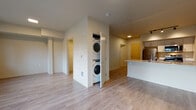 This single-floor unit offers a clean, contemporary design highlighted by light wood-style flooring that runs throughout the open living areas. The kitchen is equipped with stainless steel appliances, including a refrigerator, electric range, over-the-range microwave, and dishwasher. White cabinetry with sleek bar-pull hardware pairs with white countertops, and a peninsula with an undermount sink provides added workspace and separates the kitchen from the living area. Track lighting above the peninsula adds functional, directed illumination.

A dedicated laundry closet houses a stacked washer and dryer unit, conveniently located within the main living space. The open layout allows for flexible furniture arrangement, with the living area benefiting from generous natural light provided by multiple windows and a sliding glass door that leads to a private balcony. Outdoor views include mature trees, adding a natural element to the interior atmosphere. The bathroom features a full soaking tub with a tub-shower combo and chrome fixtures set against clean white surround panels. Overall, the space presents a functional, well-appointed design with modern finishes throughout.
