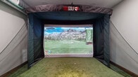 This single-floor indoor golf facility is purpose-built for an immersive simulation experience. The centerpiece is a fully enclosed TruGolf simulator bay, complete with a large high-definition projection screen, impact netting, and artificial turf hitting surface. The simulator displays detailed course data and lifelike course visuals, delivering a realistic round of golf regardless of the weather outside. Exposed industrial-style ductwork runs overhead, adding a modern utilitarian character to the space. Adjacent to the simulator bay is a comfortable lounge and viewing area, also finished with lush artificial turf flooring and featuring a bistro-style table with swivel bar stools — ideal for spectators or players between shots. A wall-mounted display screen in this area provides an additional viewing angle. The rooms are connected through rustic wood-trimmed doorways and interior windows that maintain a sense of openness while defining each functional zone. A side wall-mounted touchscreen panel offers convenient control of the simulator. Beyond the simulator area, an adjoining room is visible through the interior windows and houses fitness equipment, extending the utility of the space. Throughout, recessed lighting, clean white walls, and warm wood framing create a polished yet relaxed atmosphere well-suited for both serious practice and casual entertainment.