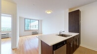 This single-floor unit showcases a clean, contemporary design throughout. The open-concept layout connects the kitchen and living area seamlessly, with warm hardwood flooring extending across both spaces to create a cohesive, inviting atmosphere. The kitchen is well-appointed with dark espresso cabinetry, stainless steel appliances including a refrigerator, microwave, and range, complemented by a large white peninsula countertop that doubles as a breakfast bar and provides additional prep space. A stainless steel sink is integrated into the peninsula, adding both style and functionality.

The living area is bright and open, benefiting from a large window that frames urban views of the surrounding cityscape, with a wall-mounted HVAC unit positioned beneath. The clean white walls throughout the unit enhance the sense of light and openness.

The bedroom is a well-proportioned, quiet space featuring the same warm hardwood floors and a walk-in closet offering generous storage. The bathroom is finished with neutral tile flooring, a dark-toned vanity with a countertop sink, a large mirror, and a wall-mounted heater, providing a functional and tidy space. Overall, the unit presents a polished, modern aesthetic with practical amenities.