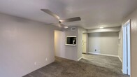 This single-floor residential unit offers a clean, functional layout with neutral grey walls and a mix of flooring throughout. The living area is carpeted and features a modern ceiling fan with integrated lighting, a front-facing window that brings in natural light, and direct access to the main entry door. A convenient pass-through window connects the living space to the kitchen, creating an open feel between the two areas. The kitchen is well-appointed with grey shaker-style cabinetry, light speckled countertops, a mosaic tile backsplash, a stainless steel sink, and black appliances including a refrigerator, range, and microwave. Wood-look vinyl plank flooring runs throughout the kitchen, adding warmth and durability. The bedrooms are carpeted and each include ceiling fans with lighting and windows with blinds that allow for natural light and privacy. A connecting doorway visible from one bedroom leads to an additional room or hallway, providing practical circulation through the unit. Transitional spaces feature a combination of carpet and vinyl plank flooring, with HVAC vents and clean white trim throughout. The overall ambiance is understated and move-in ready, with updated finishes that offer a fresh, contemporary feel.