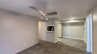 This single-floor residential unit offers a clean, functional layout with neutral grey walls and a mix of flooring throughout. The living area is carpeted and features a modern ceiling fan with integrated lighting, a front-facing window that brings in natural light, and direct access to the main entry door. A convenient pass-through window connects the living space to the kitchen, creating an open feel between the two areas. The kitchen is well-appointed with grey shaker-style cabinetry, light speckled countertops, a mosaic tile backsplash, a stainless steel sink, and black appliances including a refrigerator, range, and microwave. Wood-look vinyl plank flooring runs throughout the kitchen, adding warmth and durability. The bedrooms are carpeted and each include ceiling fans with lighting and windows with blinds that allow for natural light and privacy. A connecting doorway visible from one bedroom leads to an additional room or hallway, providing practical circulation through the unit. Transitional spaces feature a combination of carpet and vinyl plank flooring, with HVAC vents and clean white trim throughout. The overall ambiance is understated and move-in ready, with updated finishes that offer a fresh, contemporary feel.