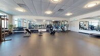 Located at 1085 Goldstream Ave in Victoria, BC, this fitness facility offers a single-level exercise room designed to support a wide range of workout routines. The space is generously sized, providing ample room for movement and equipment without feeling crowded.

The exercise room features durable rubber flooring throughout, offering both safety and comfort underfoot. A suspended drop-tile ceiling with flush-mounted lighting ensures the space is consistently and evenly illuminated. Large windows along the perimeter allow natural light to filter in, creating a bright and energizing atmosphere.

Full-length mirrored walls visually expand the room and allow users to monitor their form during workouts. The cardio section is well-appointed with treadmills, stationary bikes, a rowing machine, and elliptical trainers. The strength training area includes a multi-functional cable machine, free weights, benches, and additional resistance equipment. Stability balls, exercise mats, and a mini trampoline are also available for functional fitness and flexibility training.

A bold motivational mural featuring fitness-themed typography adds a dynamic visual accent to one wall, reinforcing the active and encouraging atmosphere of the space. A wall-mounted television provides entertainment during workouts. The facility is thoughtfully organized to accommodate both individual training and group exercise activities.