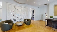 Located at 2000 Alexan Dr in Cary, NC, this single-floor community clubhouse showcases a thoughtfully designed interior that blends artistic expression with functional comfort. The entry lounge serves as an immediate focal point, featuring a striking floor-to-ceiling decorative art installation composed of colorful mixed-media elements against a crisp white backdrop. Herringbone-patterned hardwood flooring flows throughout, grounding the space with warmth and visual continuity.

The main gathering area offers generous open space with distinct seating zones. A built-in bench seat upholstered in teal, accented with vibrant yellow throw pillows, lines a bold textured blue accent wall and opens to the outdoors through full-height glass doors dressed in deep burgundy drapes. Nearby, a dramatic marble-clad fireplace wall anchors a lounge area with a wall-mounted TV and views through large windows to an outdoor patio.

A dedicated workstation nook features a built-in counter with storage cabinets, white brick tile backsplash, a golden accent ceiling, and playful pendant lighting in pastel tones. A conference-style dining area with a round wood table, modern pendant lights, and corner windows brings additional functionality to the space.

Throughout, recessed lighting, globe wall sconces, and an abundance of natural light create a bright, welcoming ambiance suited for both relaxation and productivity.