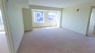 This single-floor space presents a straightforward and functional layout centered around a generously sized open room. The interior is finished with wall-to-wall plush carpeting in a neutral tone, providing a soft and comfortable underfoot experience that complements the overall simplicity of the design. The walls are painted in a light, creamy white, contributing to an airy and clean aesthetic throughout.

A pair of large windows along the far wall serves as a defining architectural feature, allowing natural light to pour into the space and illuminate the room with a bright, welcoming atmosphere. The windows also offer views of the surrounding urban exterior, adding a sense of connection to the outside environment. The ceiling is smooth and uniform, maintaining a consistent height that reinforces the open feel of the room.

The layout is notably unobstructed, creating a versatile open-plan space adaptable to a variety of uses. A subtle structural element is visible near the ceiling on one side, adding a minor architectural detail to an otherwise minimal interior. Overall, the space conveys a clean, neutral, and move-in-ready quality suited for a range of configurations.