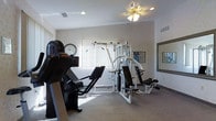 This single-floor fitness room offers a practical and well-organized space dedicated to exercise and physical wellness. The room features neutral-toned walls and light carpeted flooring, creating a clean and functional atmosphere. A ceiling fan with an integrated light fixture provides both ventilation and illumination, keeping the space comfortable during workouts.

The fitness area is equipped with a multi-station cable strength-training machine, offering a variety of resistance exercises in one compact unit. Complementing this is a cardio machine positioned toward the front of the room, providing options for aerobic training. A large wall-mounted mirror on one side enhances the sense of space while allowing users to monitor their form during exercise.

Natural light filters into the room through a window fitted with vertical blinds, brightening the space during daytime hours. A wall clock is prominently displayed, allowing users to easily track their workout duration. A posted workout reference chart near the strength equipment adds a functional touch. The overall layout is efficient, making good use of the available floor space while maintaining an open and accessible feel.