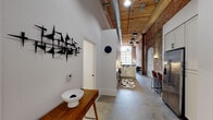 This single-floor loft in South Carolina showcases a masterful fusion of industrial character and modern sensibility. The living area immediately captures attention with dramatically tall ceilings, original exposed brick walls featuring a distinctive arched alcove, and oversized warehouse-style windows that flood the space with abundant natural light. Polished concrete floors run continuously throughout, anchoring the open layout with an understated, durable elegance.

The kitchen is thoughtfully appointed with white shaker cabinetry, granite countertops, stainless steel appliances, a matte black faucet, and a herringbone-patterned tile backsplash. Pendant lighting and an exposed ductwork system overhead reinforce the loft's industrial aesthetic while maintaining practicality.

A wood-plank ceiling with heavy timber beams adds warmth to the elongated corridor connecting the living spaces to the private quarters. The bedrooms feature soaring white walls, original structural columns, and polished concrete floors, creating a calm, gallery-like atmosphere. One bedroom benefits from an original exposed brick column as a striking architectural accent.

The bathroom is finished with subway tile, a glass-enclosed shower, dark cabinetry with granite countertops, and matte black fixtures throughout. A dedicated laundry room provides full-size washer and dryer units with built-in shelving, adding practical convenience to this characterful space.