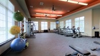 This fitness center offers a spacious, single-floor layout designed to accommodate a full range of workout activities. The room features a distinctive tray ceiling finished in a warm terracotta tone, contrasted by neutral beige walls, creating an energizing yet welcoming atmosphere. A ceiling fan centered within the tray ceiling aids in air circulation throughout the space. Rows of cardio equipment, including treadmills and elliptical machines, are aligned along one side of the room, positioned to take advantage of the natural light streaming in through the numerous windows fitted with horizontal blinds. A wall-mounted flat-screen television provides entertainment during workouts. On the opposite side, strength training equipment, resistance machines, and free weights such as stability balls and medicine balls are available for varied exercise routines. The flooring consists of durable, low-pile carpet suitable for both cardio and strength activities. Recessed lighting and overhead fluorescent fixtures ensure the space is well-lit at all hours. The overall layout offers generous open floor space, allowing for comfortable movement between equipment stations.