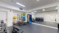 Located at 8732 Town and Country Blvd in Ellicott City, Maryland, this dedicated fitness room offers a compact yet highly functional workout environment. The space features durable rubber flooring throughout, providing excellent traction and cushioning for a variety of exercises. Bright recessed lighting and flush rectangular light panels illuminate the room evenly, creating a clean, energetic atmosphere.

A defining feature of the space is the large floor-to-ceiling mirror spanning an entire wall, which visually expands the room while allowing users to monitor their form during workouts. Opposite the mirror, wall-mounted racks display a colorful array of stability and medicine balls, adding both functionality and visual vibrancy to the space.

The fitness room is well-stocked with strength training equipment, including a multi-tiered dumbbell rack with a full range of weights, kettlebells, an adjustable workout bench, and resistance bands. A freestanding ball storage tower and additional fitness accessories round out the available equipment.

Natural light pours in through a striking wall of white-framed glass doors and transom windows on one side of the room, offering views of an outdoor patio area and pool surroundings. These doors also provide direct access to the exterior, seamlessly connecting indoor and outdoor amenities. The overall design is clean and functional, making this fitness room a practical and inviting space for daily exercise.