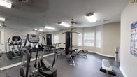 This fitness center offers a comprehensive workout environment within a single, thoughtfully arranged space. The room is outfitted with dark rubber flooring throughout, providing a durable and functional surface suited for a variety of exercise activities. A ceiling fan and recessed fluorescent lighting ensure the space remains well-lit and comfortable during use.

Cardio equipment is a focal point, featuring multiple True-brand treadmills, elliptical trainers, and a stationary bike, each positioned on protective floor mats. A wall-mounted flat-screen television is conveniently placed to keep users entertained during cardio sessions. A multi-station cable strength machine anchors the strength-training area, accompanied by adjustable benches and additional resistance equipment for a full-body workout.

Large wall mirrors span two sides of the room, visually expanding the space and allowing users to monitor their form. A bay window arrangement on one wall admits natural light, contributing to an open, airy atmosphere. Instructional fitness and stretching posters are displayed on the walls, offering guidance for users of all experience levels. A sanitizing station is also present, supporting cleanliness throughout the space.