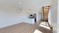 This two-story space showcases a contemporary, minimalist design characterized by clean white walls, light hardwood flooring, and an abundance of natural light streaming across the open-plan main floor. The living area is generously proportioned, offering a versatile layout that flows seamlessly into the kitchen zone.

The kitchen features a sleek white island with an integrated sink, complemented by white upper cabinetry and a suite of stainless steel appliances, including a refrigerator, microwave, and gas range. Pendant lighting hangs above the island, adding a refined touch to the functional space. Recessed ceiling lights throughout provide additional illumination.

A wall-mounted mini-split HVAC unit ensures efficient climate control across the main living area. A standout architectural feature is the open-tread staircase constructed with warm-toned wood slats, providing an elegant visual connection between the two floors while allowing light to pass freely through.

The overall ambiance is bright, airy, and modern, with a neutral palette that creates a versatile and welcoming environment throughout.