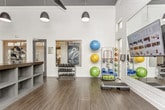 This modern fitness center is thoughtfully designed to accommodate a full range of workout activities across a single open floor. The space is divided into distinct functional zones, each purpose-built for a specific type of training. The cardio area features a lineup of TRUE-brand upright and recumbent bikes, treadmills, and elliptical machines, all positioned beside expansive floor-to-ceiling windows that invite generous natural light and offer views of the surrounding landscaped exterior. A dedicated strength training zone houses a multi-station cable machine and benches, set atop patterned blue-accent carpet that visually delineates the area. A central island countertop with open shelving provides convenient storage and organization at the heart of the space. The functional training area features hardwood-style flooring and is outfitted with a dumbbell rack, resistance bands, stability and medicine balls, yoga mats, and step platforms, alongside a large wall-mounted display screen for guided workouts. Full-length mirrors amplify the sense of space and assist with form. Dark pendant lights hang from the elevated ceilings, complemented by ceiling fans that maintain air circulation. The overall aesthetic is clean, contemporary, and bright, creating an energizing environment conducive to focused exercise.
