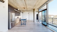Located in Hollywood, this 2-bedroom, 2-bathroom residence showcases a striking industrial-modern aesthetic that blends raw architectural elements with refined finishes. The open-plan living and kitchen area is anchored by an exposed concrete ceiling with visible ductwork and structural columns, creating an urban loft ambiance. Light hardwood flooring flows throughout, complementing the bright, airy atmosphere generated by expansive floor-to-ceiling windows and sliding glass doors that open to a balcony with sweeping views of the Hollywood Hills and surrounding cityscape.

The kitchen is thoughtfully designed with dual-tone cabinetry — dark upper cabinets contrasting with light wood lower units — paired with a white tile backsplash, stainless steel appliances, and a freestanding island work surface. A hallway connects the living spaces to the private quarters, where a stacked in-unit washer and dryer is conveniently housed. The spacious primary bedroom features a sliding barn-style door leading to a built-in closet and direct access to an en suite bathroom with a tiled soaking tub. The second bedroom also includes a built-in closet with exposed concrete column detailing. Bathrooms are finished with large-format floor tiles, white subway tile surrounds, and sleek matte black hardware and open shelving.