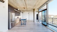 Located in Hollywood, this 2-bedroom, 2-bathroom residence showcases a striking industrial-modern aesthetic that blends raw architectural elements with refined finishes. The open-plan living and kitchen area is anchored by an exposed concrete ceiling with visible ductwork and structural columns, creating an urban loft ambiance. Light hardwood flooring flows throughout, complementing the bright, airy atmosphere generated by expansive floor-to-ceiling windows and sliding glass doors that open to a balcony with sweeping views of the Hollywood Hills and surrounding cityscape.

The kitchen is thoughtfully designed with dual-tone cabinetry — dark upper cabinets contrasting with light wood lower units — paired with a white tile backsplash, stainless steel appliances, and a freestanding island work surface. A hallway connects the living spaces to the private quarters, where a stacked in-unit washer and dryer is conveniently housed. The spacious primary bedroom features a sliding barn-style door leading to a built-in closet and direct access to an en suite bathroom with a tiled soaking tub. The second bedroom also includes a built-in closet with exposed concrete column detailing. Bathrooms are finished with large-format floor tiles, white subway tile surrounds, and sleek matte black hardware and open shelving.