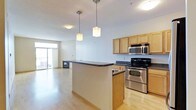 This single-floor space offers a thoughtfully designed open-concept layout that seamlessly connects the kitchen and living areas. The kitchen is a standout feature, outfitted with stainless steel appliances including an electric range and built-in microwave, light wood cabinetry providing ample storage, and a dark-topped peninsula that serves as both a workspace and a natural divider between the kitchen and the adjoining living area. Pendant lights with warm-toned shades hang above the peninsula, adding a welcoming ambiance to the space. The flooring throughout is a light hardwood, contributing to a bright and airy feel. The living area benefits from generous natural light streaming in through large windows, which also offer views to the outdoors. The neutral wall tones and clean architectural lines lend the space a contemporary, understated aesthetic. Flush-mount ceiling fixtures provide additional overhead lighting throughout. Overall, the space balances functional design with a modern sensibility, making it a practical and comfortable environment.