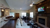 This single-story space showcases an open-concept layout that seamlessly connects the living and dining areas, creating a cohesive and functional environment. The living room is anchored by a tiled fireplace framed with rich, dark wood columns and a substantial mantle, providing a striking focal point. A ceiling fan with a light fixture is mounted at the apex of the vaulted ceiling, which adds a sense of spaciousness and volume to the room. Recessed lighting complements the natural light streaming in through multiple double-hung windows fitted with horizontal blinds.

The living area is furnished with a plush, light-colored sectional sofa and a dark-framed glass coffee table, set atop a patterned area rug laid over dark hardwood flooring that extends throughout the space. Transitioning naturally from the living room, the dining area accommodates a dark wood dining table surrounded by upholstered chairs. A large wooden armoire adds storage and character near the dining space. Lush green plants provide organic touches, enhancing the overall warm and inviting ambiance of this well-appointed, single-level home.