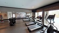 This fitness center is thoughtfully designed to support a comprehensive workout experience within a single-floor layout. The space is finished with dark rubberized flooring throughout, providing both durability and comfort underfoot, while neutral gray walls and recessed lighting create a clean, focused atmosphere.

The cardio zone is prominently featured along one wall, housing multiple treadmills and elliptical cross-trainers arranged in a row. Large windows along this side of the room flood the space with natural light and offer views of the outdoor surroundings, making workouts feel open and connected to the exterior environment. A wall-to-wall mirror runs along an adjacent wall, visually expanding the space and allowing users to monitor their form.

A dedicated free weight area includes a full rack of dumbbells, while a cable and strength training machine provides options for resistance-based exercise. A leg press and weight bench add further versatility for strength training routines. A medicine ball rack stocked with a variety of weighted balls rounds out the functional fitness equipment.

Motivational typography is displayed prominently on one accent wall, featuring phrases designed to inspire and energize users during their workouts. Roman shade window treatments add a decorative touch. The space also includes a wall-mounted television, enhancing the cardio experience. The overall ambiance is modern, energetic, and purposefully designed for an effective fitness environment.
