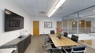 This single-floor professional office space is designed with functionality and modern aesthetics in mind. The centerpiece of the space is a polished conference room featuring a clean-lined wooden conference table surrounded by ergonomic black leather chairs, creating an inviting yet professional atmosphere for meetings and collaboration. A large flat-screen display is mounted on one wall, offering seamless presentation capabilities, while a credenza beneath provides additional storage and surface space.

The room is illuminated by recessed linear fluorescent lighting set within a drop-tile ceiling, ensuring bright, even lighting throughout. Warm wood-toned doors add a natural contrast to the crisp white walls, one of which is adorned with a framed piece of artwork that adds a touch of character to the space. A vase of fresh pink orchids on the conference table introduces a subtle organic element.

Floor-to-ceiling glass partition walls on one side of the conference room provide visual connectivity to the adjacent workspace, maintaining an open and transparent feel while preserving acoustic separation. Patterned commercial carpeting grounds the space with a practical, durable finish. Overall, this office exudes a clean, contemporary, and professional ambiance well-suited for a dynamic business environment.
